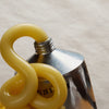 A close-up of a thick, glossy yellow substance, likely a thick cream or gel, draped over the open neck of a silver metal squeeze tube against a warm brown gradient background.