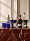 A collection of bar accessories arranged on a reddish-brown marble block against a cream paneled wall. The set includes cobalt blue and emerald green martini glasses, a silver shaker, and several liquor bottles.