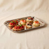 A rectangular silver metal tray with a decorative beaded border, resting on a draped cream fabric background.