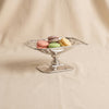 A polished silver pedestal bowl with a square rim, set against a draped beige fabric background.