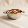 A small polished stainless steel bowl with two decorative shell-shaped side handles, photographed on a soft beige fabric background.