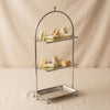 A three-tiered metal serving cart with a polished silver finish, featuring a curved top frame and three rectangular trays, set against a draped beige fabric backdrop.