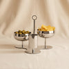A polished silver metal three-piece serving set on a draped beige fabric background, featuring two stemmed bowls and a central cylindrical holder with a top loop handle.