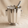 A polished silver fluted ice bucket with a scalloped rim and two hanging loop handles, set against a draped beige fabric background.