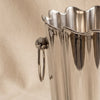 A polished silver fluted ice bucket with a scalloped rim and two hanging loop handles, set against a draped beige fabric background.