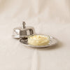 A small silver-toned metal butter dish with a matching lid and a clear glass inset dish, photographed on a soft beige fabric background.