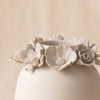 A matte ceramic vase with a rounded body and a rim decorated with sculpted flowers, shown on a draped beige fabric background.
