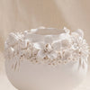 A white ceramic planter with a wide, rounded base and a decorative rim featuring sculpted white flowers and leaves, set against a draped beige fabric background.